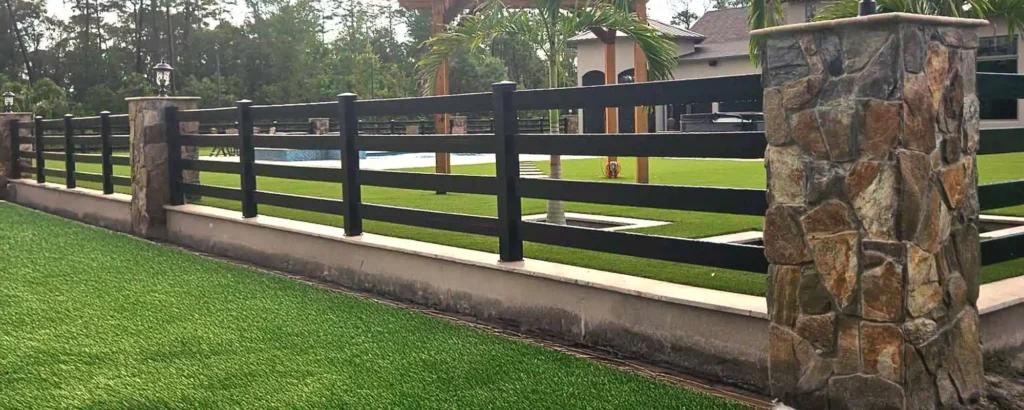 Black four-rail vinyl fence between stone pillars beside a lawn.