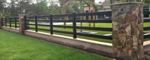 Black four-rail vinyl fence between stone pillars beside a lawn.